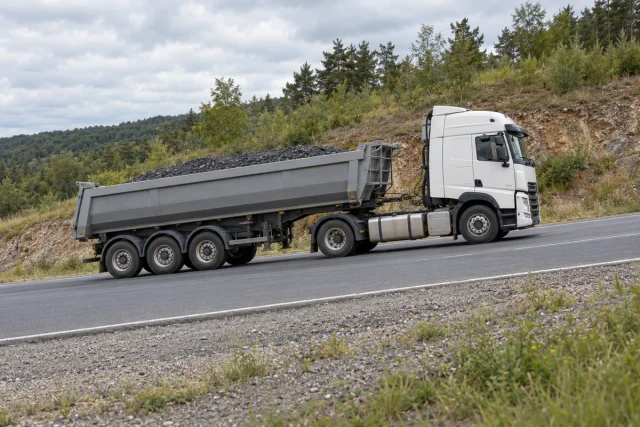 Caminhão rodoviário articulado com semirreboque basculante transportando material mineral em uma estrada pavimentada em aclive.