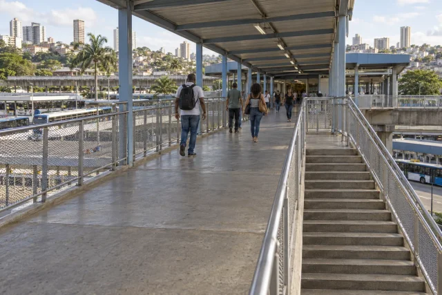 Usuários caminham por passarela coberta com escada e guarda-corpos, junto a plataformas e ônibus de transporte coletivo.