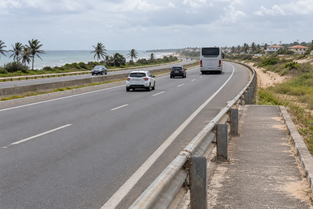 Ônibus e automóveis em rodovia costeira de pistas separadas, com mar distante e guardrail em primeiro plano.
