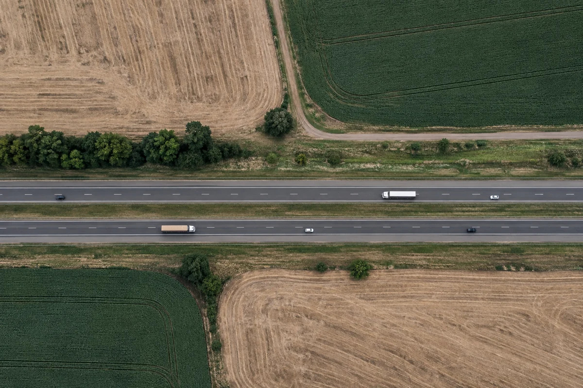 Vista vertical de rodovia de pistas separadas atravessando parcelas agrícolas de cores e texturas distintas.