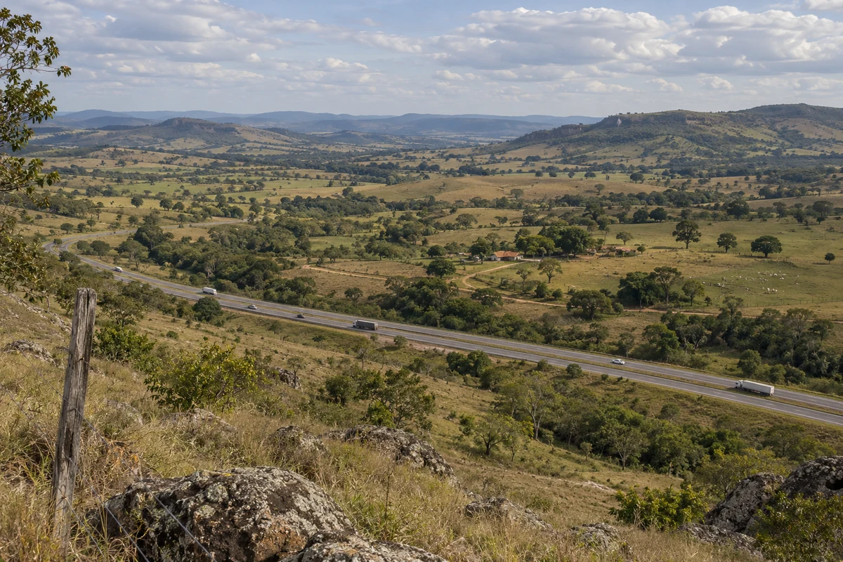 Rodovia atravessando vale rural amplo, com campos, pequenas edificações e veículos distantes.