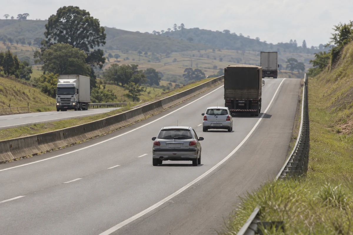 Caminhões e automóveis em rodovia com rampa suave, vistos em perspectiva comprimida junto ao guardrail.
