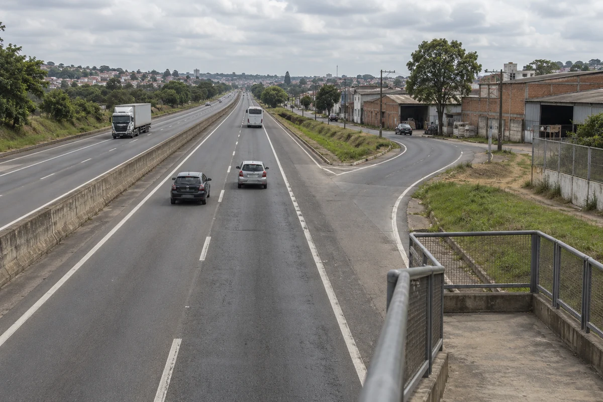 Rodovia de pistas separadas junto a galpões urbanos, com acesso lateral e tráfego esparso.