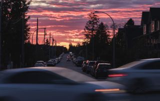 Uma rua cheia de muito tráfego sob um céu colorido ao entardecer ou pôr do sol, com carros dirigindo pela rua e alguns prédios ao lado (uma foto com efeito de inclinação e deslocamento de 0,159).