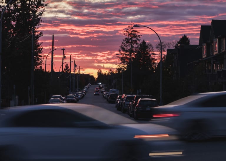 Uma rua cheia de muito tráfego sob um céu colorido ao entardecer ou pôr do sol, com carros dirigindo pela rua e alguns prédios ao lado (uma foto com efeito de inclinação e deslocamento de 0,159).