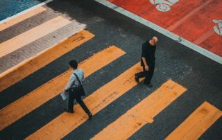 Dois homens caminhando por uma faixa de pedestres na cidade, um deles está carregando uma bolsa e o outro está carregando uma mochila e uma bolsa. (uma foto de inclinação: 0,207)