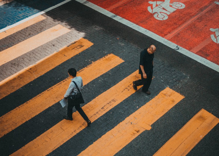 Dois homens caminhando por uma faixa de pedestres na cidade, um deles está carregando uma bolsa e o outro está carregando uma mochila e uma bolsa. (uma foto de inclinação: 0,207)