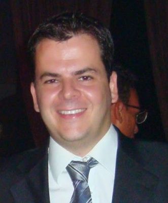 Um homem de terno e gravata sorrindo para uma foto em uma sala escura com outras pessoas ao fundo e uma mulher sorrindo ao fundo. (Um retrato de personagem: 0,284)