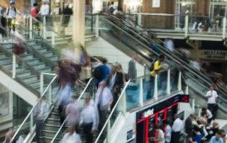 Um grupo de pessoas andando para cima e para baixo em um conjunto de escadas em um prédio com uma escadaria que leva a uma área de segundo andar, com um segundo piso, (uma foto em tilt shift: 0,241).