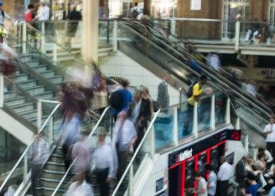 Um grupo de pessoas andando para cima e para baixo em um conjunto de escadas em um prédio com uma escadaria que leva a uma área de segundo andar, com um segundo piso, (uma foto em tilt shift: 0,241).