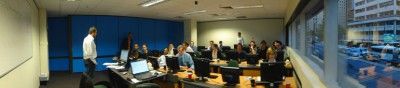 Um grupo de pessoas em pé em um escritório com uma grande janela olhando para a cidade do lado de fora do prédio, e um homem em pé em um computador, (uma foto colorida: 0.452).