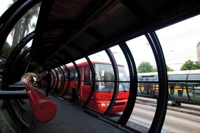 Um ônibus vermelho está estacionado em um túnel ao lado de uma calçada com um bonde vermelho e uma cerca vermelha e uma linha amarela. (uma pintura mate detalhada:0.186)