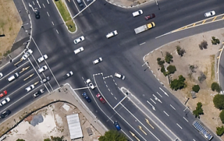 Uma vista aérea de um cruzamento movimentado com carros e caminhões em seus lados e uma rua com uma luz amarela no topo do cruzamento, (uma fotografia tilt shift: 0,275).