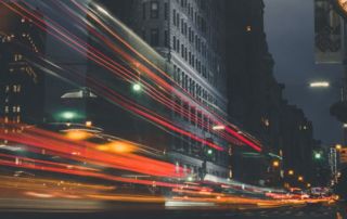 Um relógio em um poste em uma cidade à noite com longa exposição do tráfego e edifícios ao fundo com luzes riscando pelo relógio. (uma foto: 0,094)