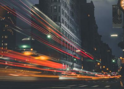 Um relógio em um poste em uma cidade à noite com longa exposição do tráfego e edifícios ao fundo com luzes riscando pelo relógio. (uma foto: 0,094)