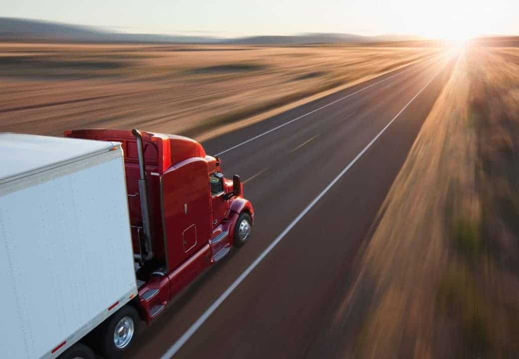 Um caminhão semi vermelho dirigindo em uma estrada ao lado de um campo de grama e árvores ao pôr do sol ou nascer do sol, com o sol brilhando no horizonte. (uma foto de estoque: 0,447)