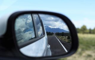 O espelho lateral de um carro reflete uma cadeia de montanhas no espelho retrovisor de um carro em uma estrada no campo. No espelho de vista lateral, há um carro (uma imagem de estoque: 0,496).