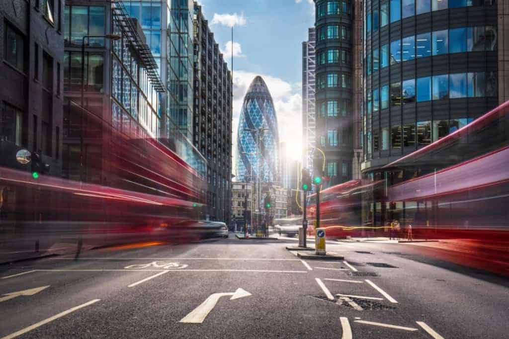 Uma rua da cidade com ônibus de dois andares passando ao longe e prédios ao fundo com um céu azul e algumas nuvens no fundo (uma foto de estoque: 0,337).