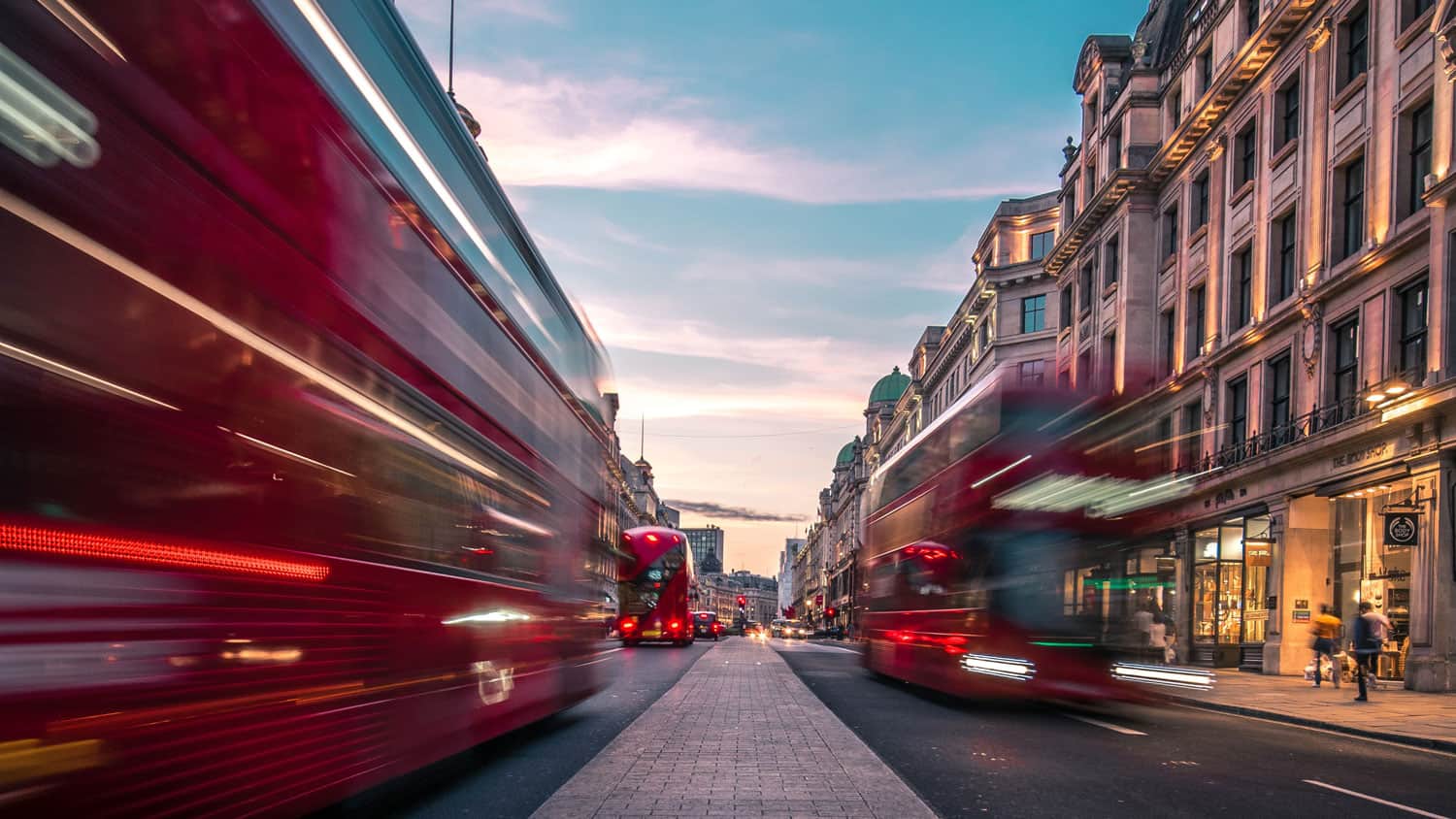 Dois ônibus de dois andares dirigindo por uma rua ao lado de prédios altos com luzes de trânsito neles e uma pessoa caminhando na calçada perto da rua com um ônibus vermelho de dois andares, (uma foto de estoque: 0.258)