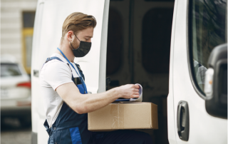 Um homem com uma caixa e uma máscara no rosto está carregando uma van com uma caixa de papelão e outra caixa de papelão do lado da van. (uma foto de estoque: 0,566)