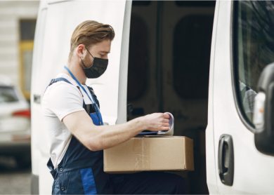 Um homem com uma caixa e uma máscara no rosto está carregando uma van com uma caixa de papelão e outra caixa de papelão do lado da van. (uma foto de estoque: 0,566)