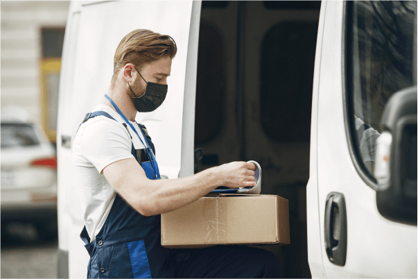 Um homem com uma caixa e uma máscara no rosto está carregando uma van com uma caixa de papelão e outra caixa de papelão do lado da van. (uma foto de estoque: 0,566)