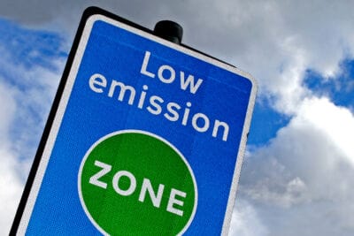 um cartaz azul que diz zona de baixa emissão com um fundo de céu e nuvens ao fundo com um círculo verde que diz zona de baixa emissão. (um cartaz: 0,233)